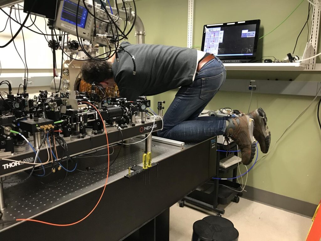 Ben kneeling uncomfortably on an optical table and struggling with some uncooperative cryostat hardware.