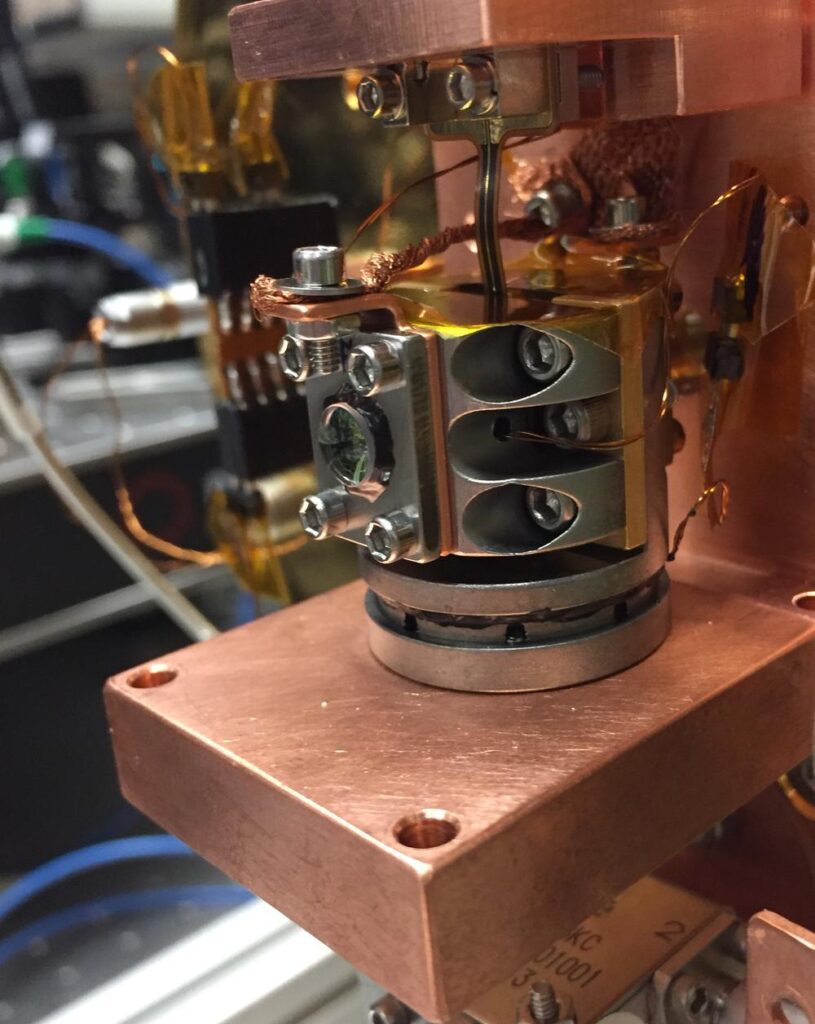 The fully assembled electro-optic transducer, enclosed in a 1" cylinder made of an invar alloy, mounted on a copper plate. A lens to couple a laser beam into the optical cavity and electrical wiring to couple signals into the superconducting circuit are visible.