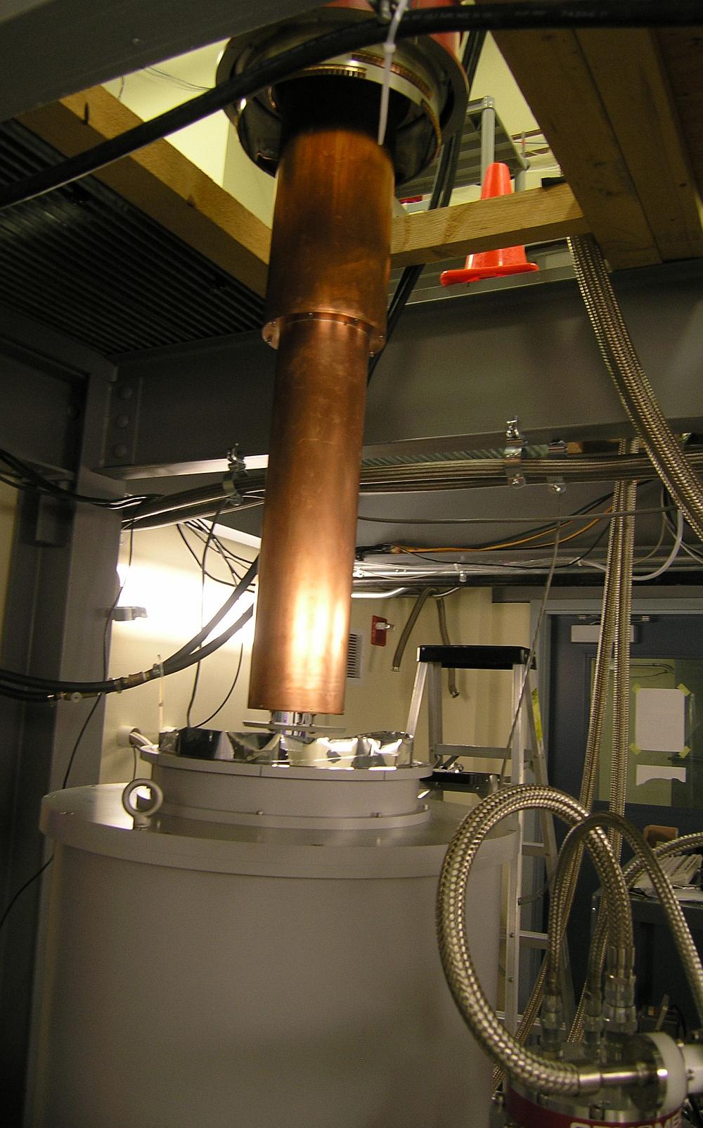 The cavity assembly, encased in a copper thermal shield, hangs from the dilution refrigerator, which has been hoisted into the air and positioned above the magnet bore for insertion.