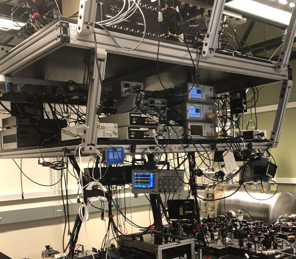 A photo of my postdoc lab: large shelves hang from the ceiling above an optical table on which are mounted lenses and mirrors and other equipment for routing and manipulating laser beams. The shelves are filled with electronic equipment, with cables running all over the place.