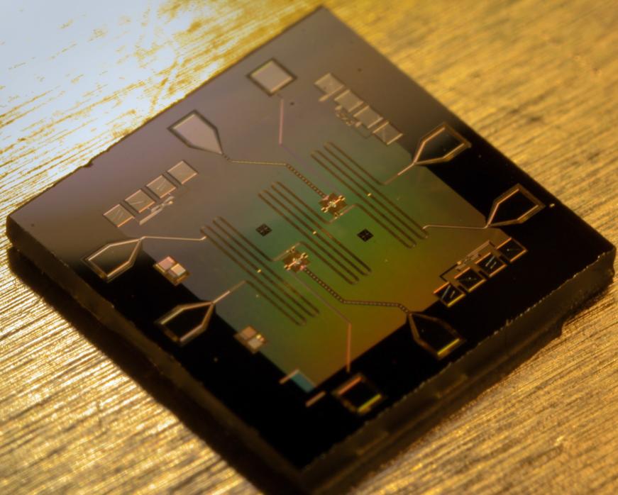 A square chip, roughly a quarter inch along either side, made of synthetic sapphire, and patterned with strips of superconducting metal to form qubits and other resonant circuits. The two qubits on the chip each occupy about 1% of its surface area. Large pads at the edge of the chip, connected to the qubits and resonators through superconducting transmission lines, can be wired up to a conventional circuit board for control and measurement.